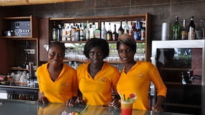 Three individuals in yellow shirts standing behind a bar with various bottles and glasses. - Leo's Beach Hotel & Restaurant - Adults Only (Brufut)