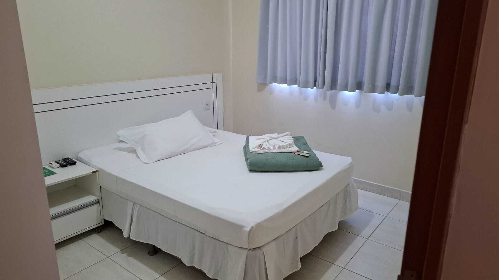 A neatly made bed with a white bedspread and a green pillow, a bedside table with a phone and a remote control, and a window with white curtains.