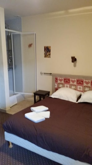Una habitación de hotel con cama, mesita de noche, luz empotrada en la pared y puerta que da al baño.