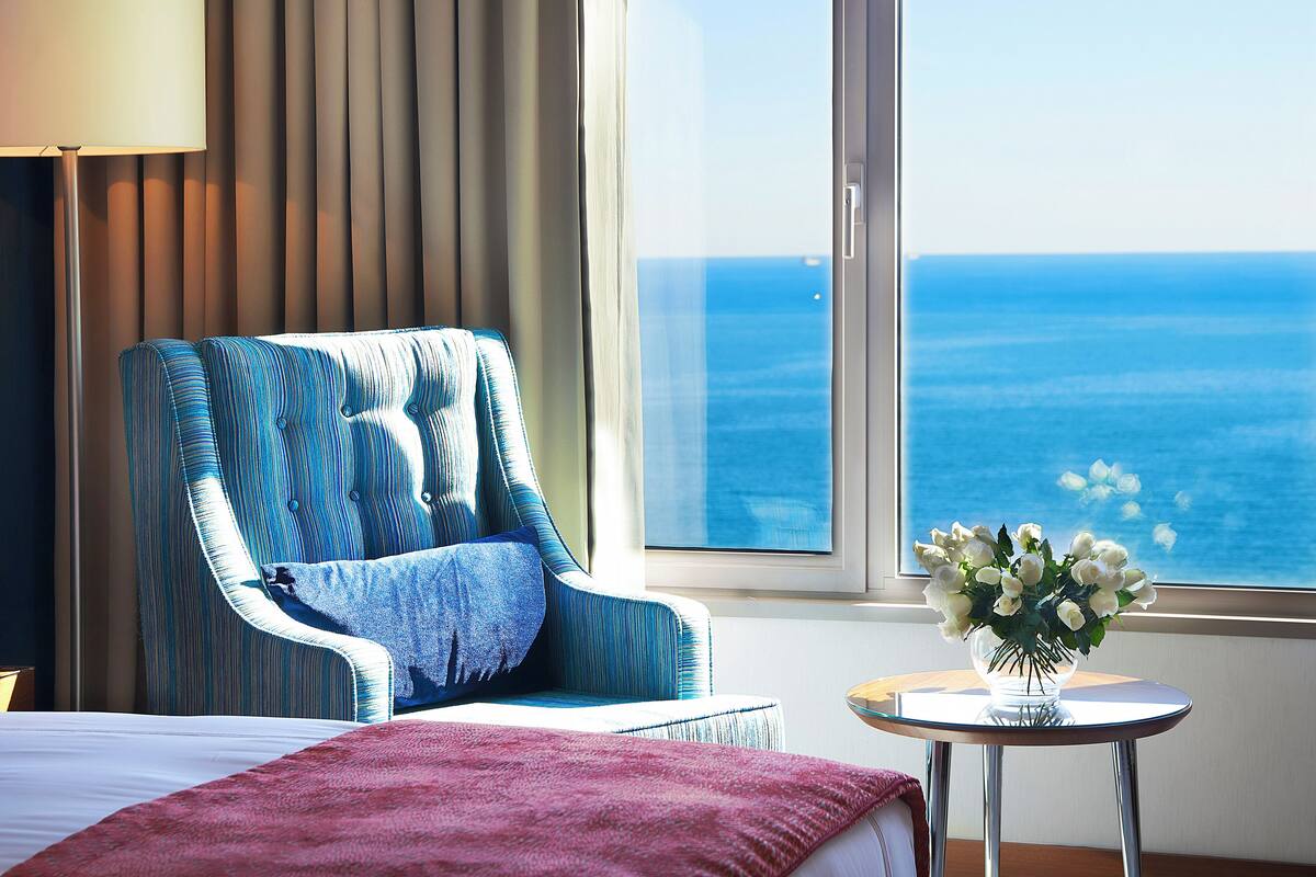 A hotel room with a large window offering a view of the ocean, a bed, a chair with a patterned cushion, a small table with a vase of flowers, and a lamp.