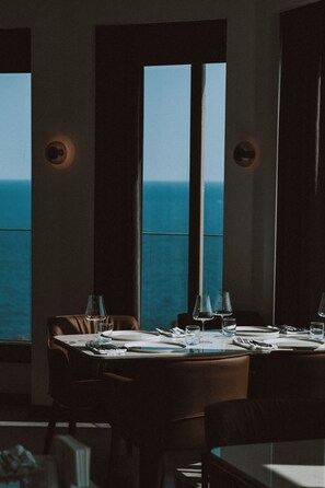Mesa de comedor para dos con vista al océano a través de amplios ventanales.
