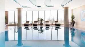 An indoor swimming pool with clear glass walls, sun loungers, and large windows offering a view of trees. - Divan Mersin (Mersin)