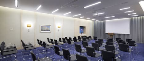 Een vergaderzaal met rijen zwarte stoelen, een blauw tapijt, een projectiescherm en een spreekgestoelte.