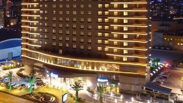 A multi-story hotel with a prominent "divan" sign on the top. It features a curved facade with evenly spaced windows and a well-lit entrance area with palm trees and a fountain. - Divan Mersin (Mersin)