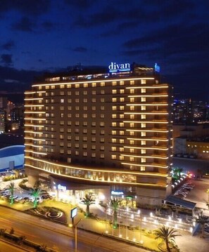 A multi-story hotel with a prominent "divan" sign on the top. It features a curved facade with evenly spaced windows and a well-lit entrance area with palm trees and a fountain. - Divan Mersin (Mersin)