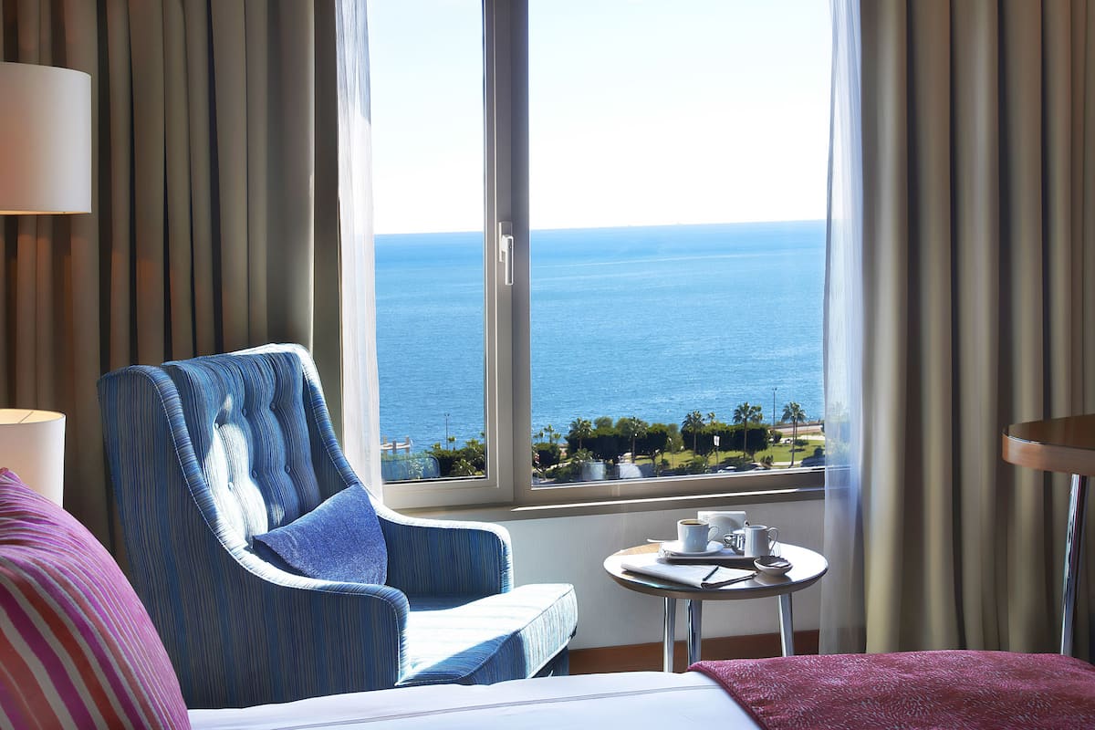 A hotel room with a large window offering a view of the sea, a bed, a chair, a small table with a cup and saucer, and a lamp.