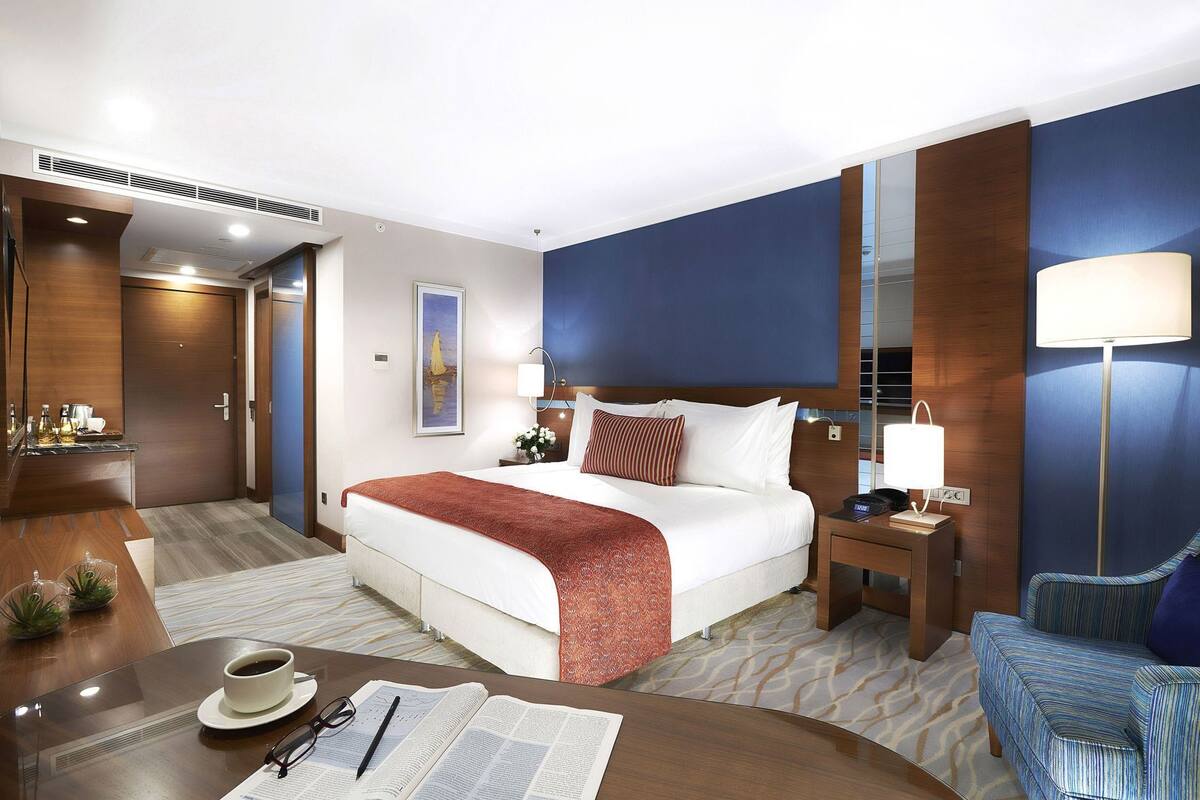 A hotel room with a bed, bedside table, lamp, chair, and a desk with a cup and glasses.