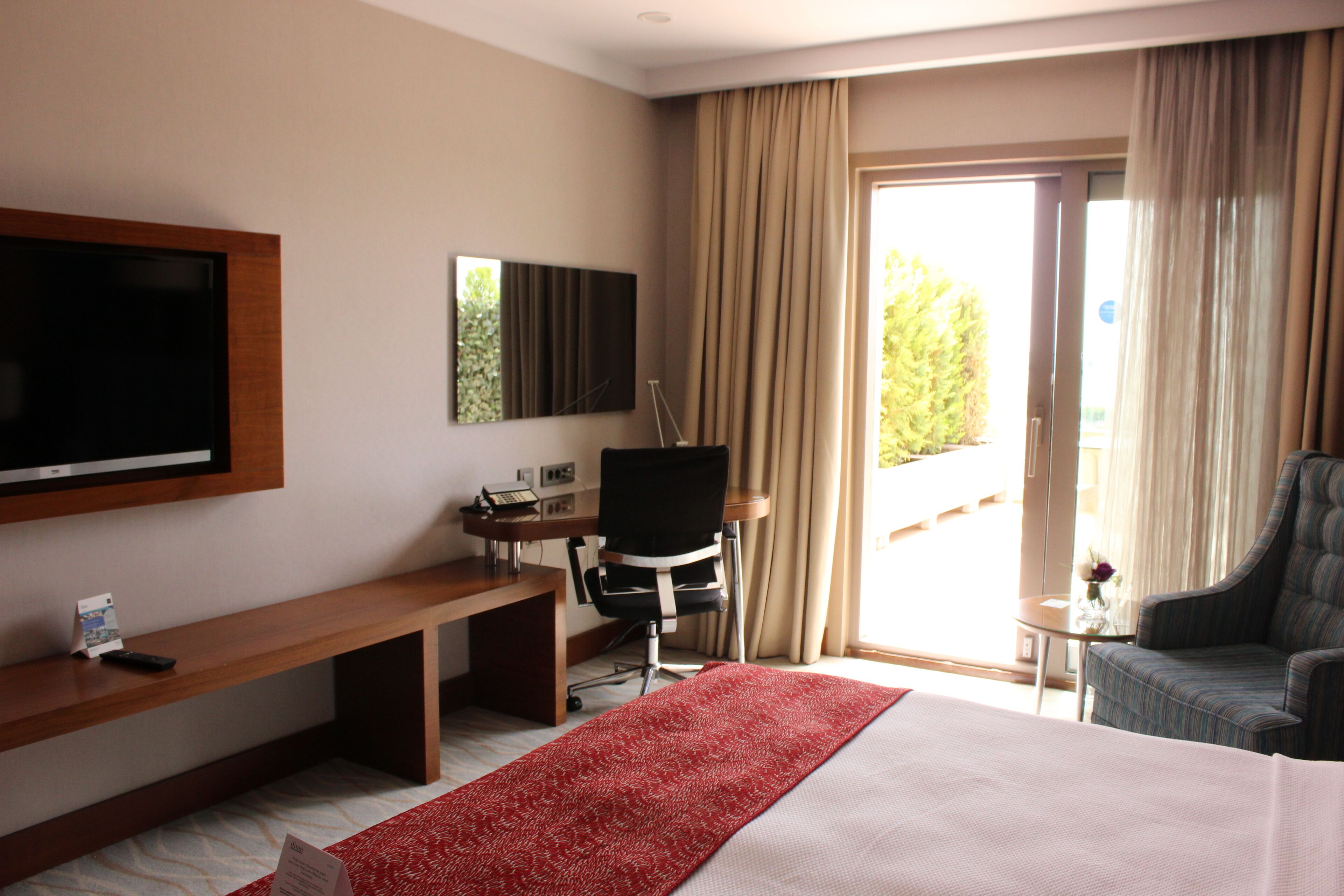 Habitación de hotel con una cama grande, un escritorio con silla, televisor montado en la pared y un balcón con cortinas.