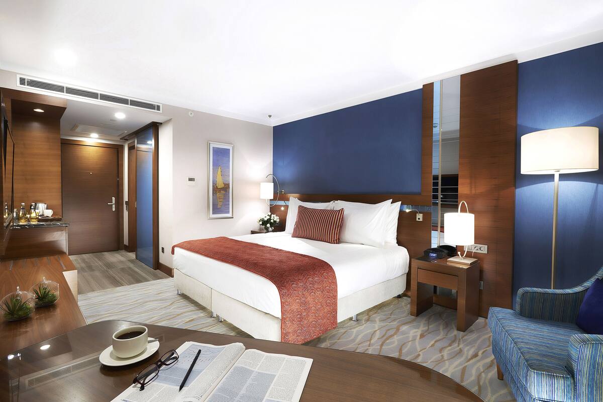 A hotel room with a bed, a desk with a cup and glasses, a chair, a nightstand with a lamp, and a large mirror.