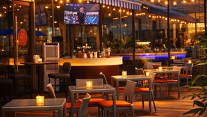Zona de bar al aire libre con mobiliario de mimbre, velas y una pantalla de televisión mostrando un evento deportivo.