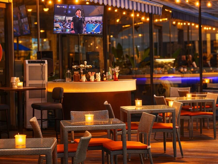 Área de bar ao ar livre com mobiliário de vime, velas e uma televisão exibindo um evento esportivo.