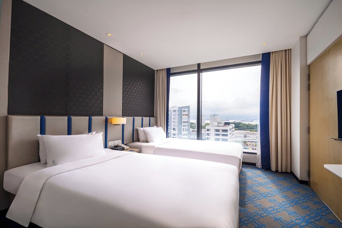 Ein Hotelzimmer mit zwei Betten, einem großen Fenster mit Blick auf die Stadt, einem Flachbildfernseher und einer Minibar.