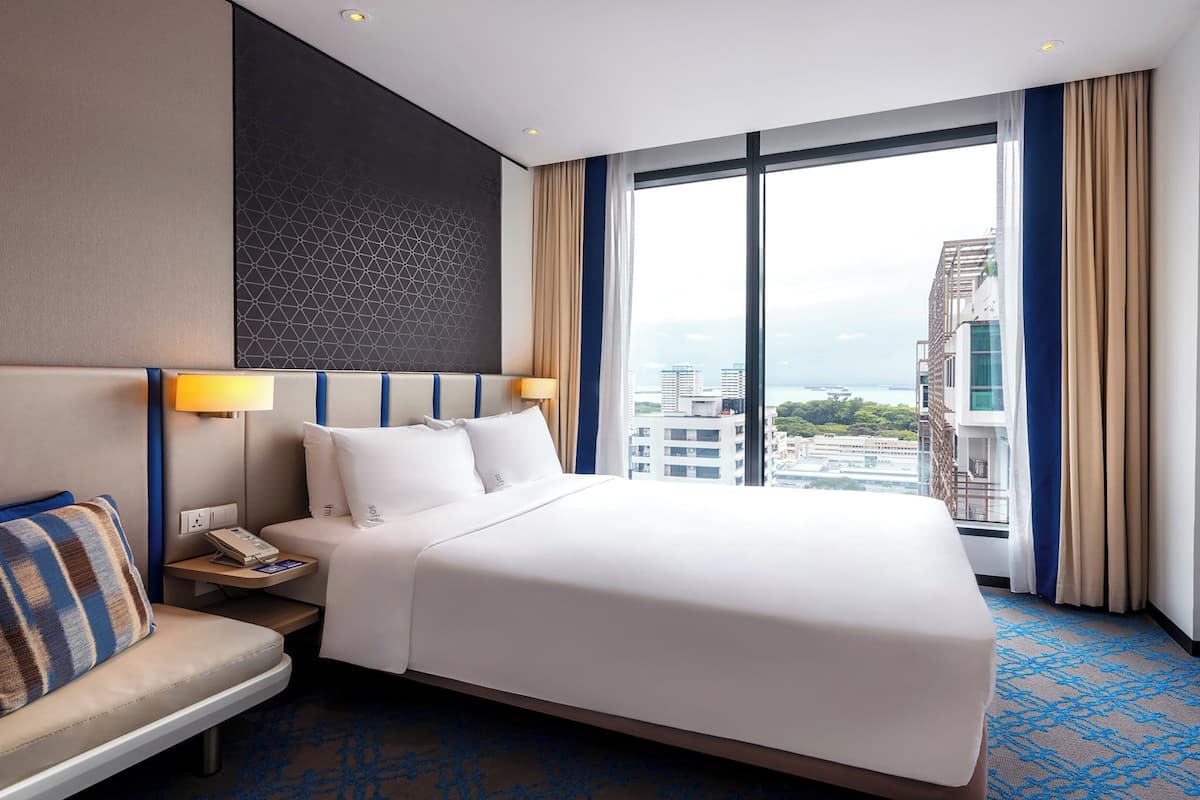 Een hotelkamer met een groot bed, een nachtkastje met een lamp, een stoel en een uitzicht op de stad door grote ramen.
