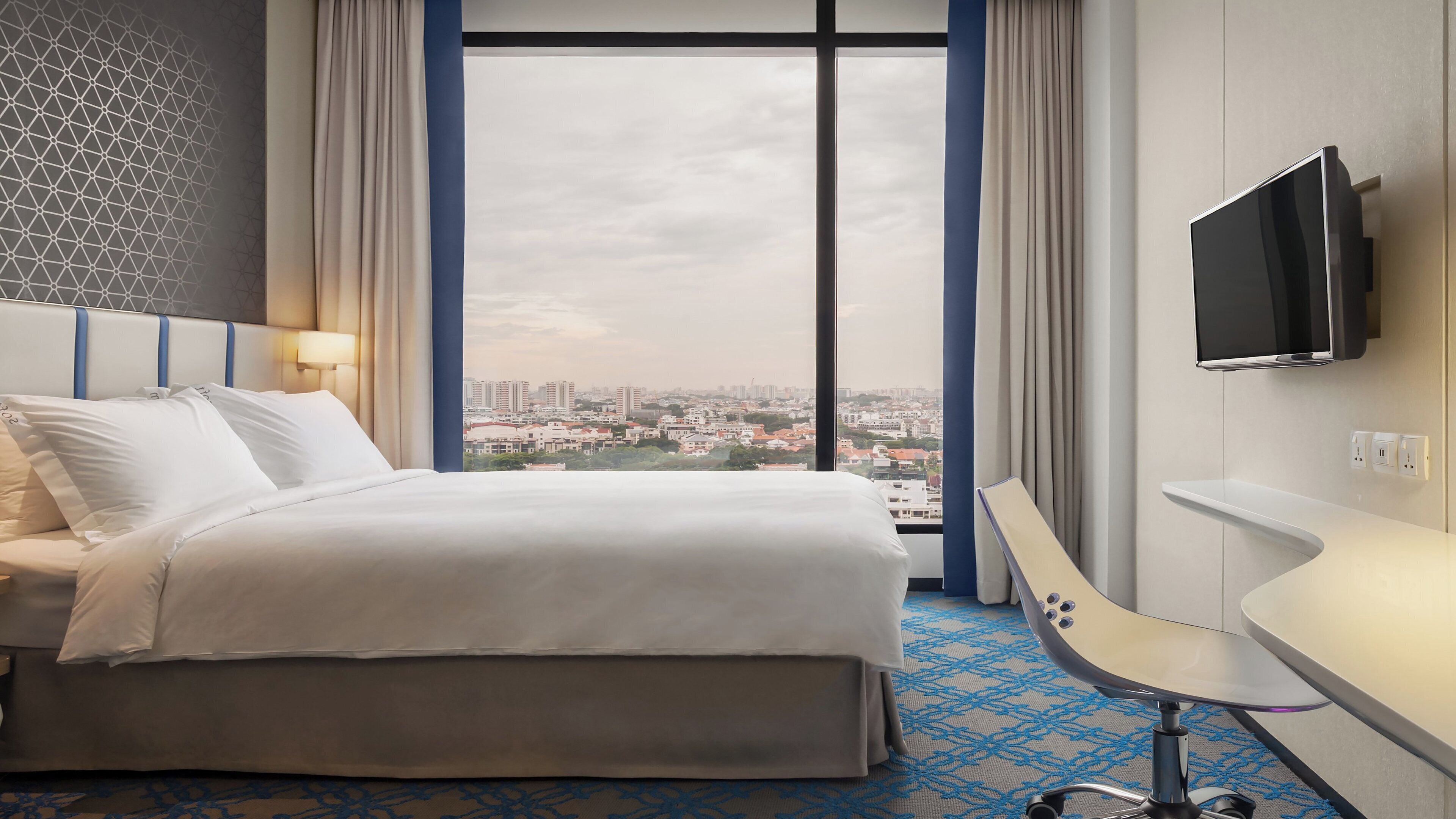 A hotel room with a bed, a desk, a TV, and a large window with a city view.