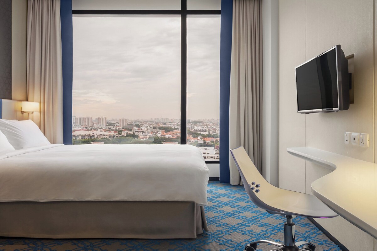Ein Hotelzimmer mit einem Bett, einem Schreibtisch, einem Fernseher und einem großen Fenster mit Blick auf die Stadt.