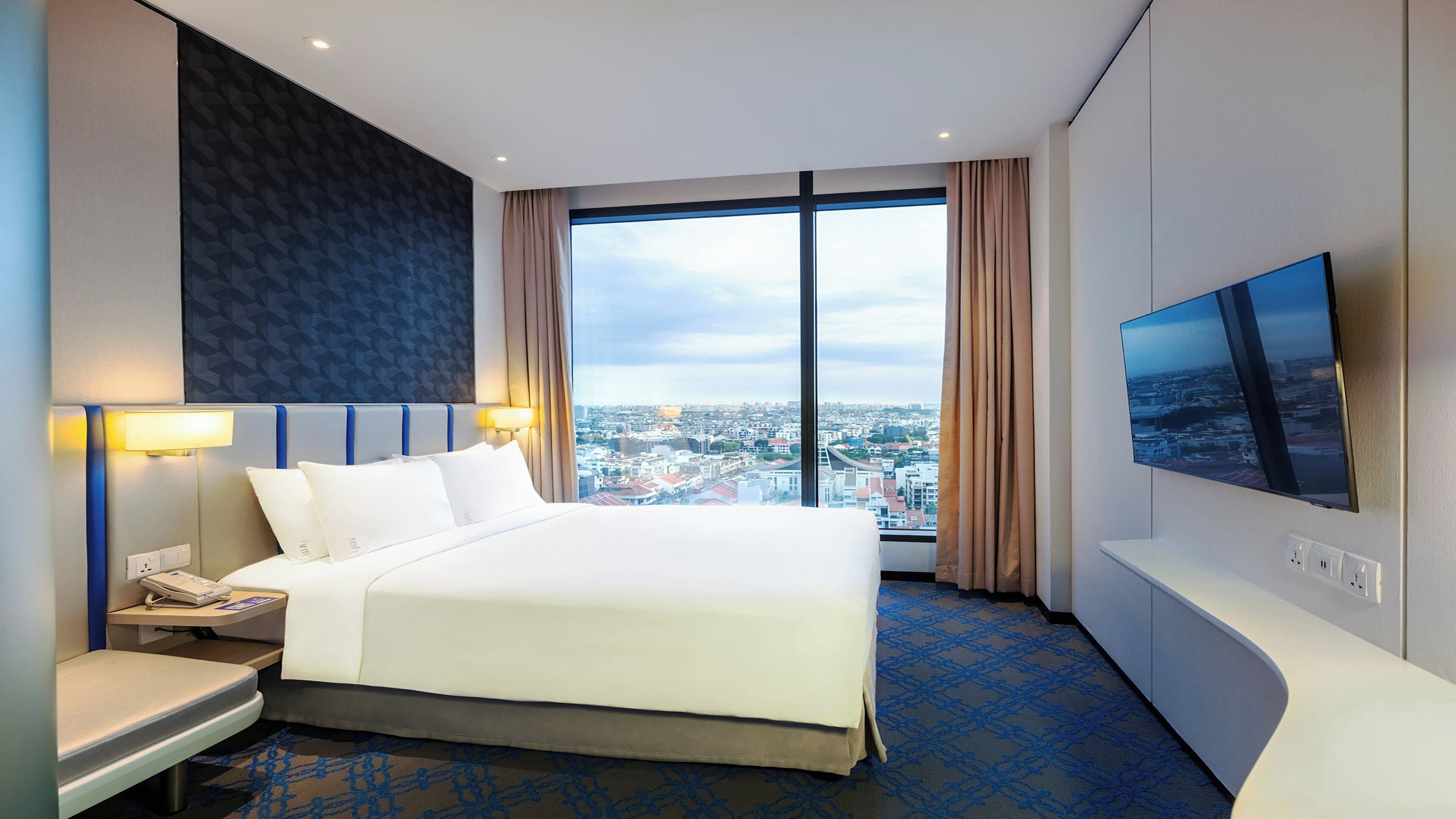 Habitación de hotel con una cama grande, un televisor en la pared, un escritorio con teléfono y vistas a la ciudad a través de amplias ventanas.