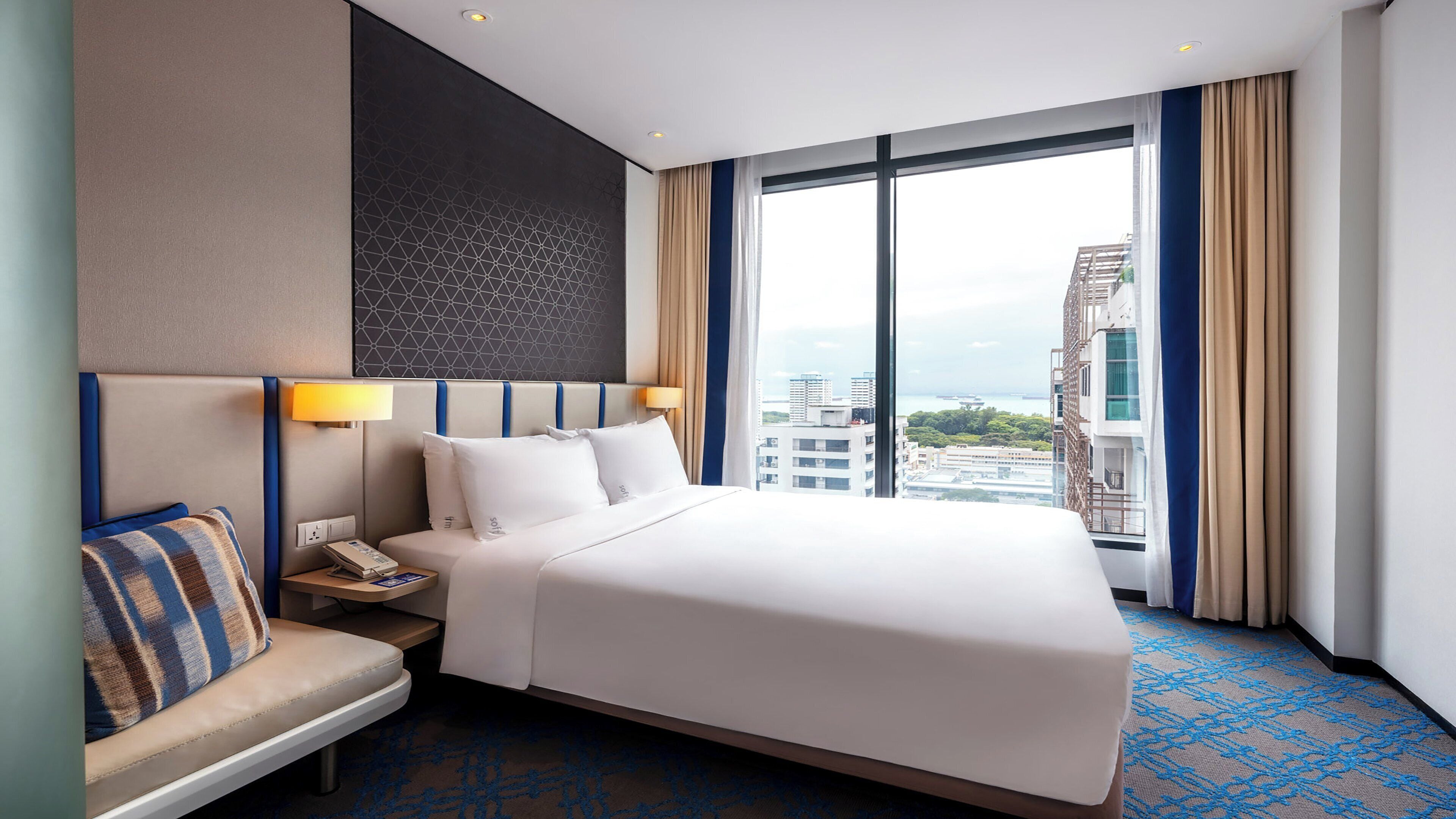 Habitación de hotel con una cama grande, una mesita de noche con lámpara, un área de descanso y vistas de la ciudad a través de amplias ventanas.