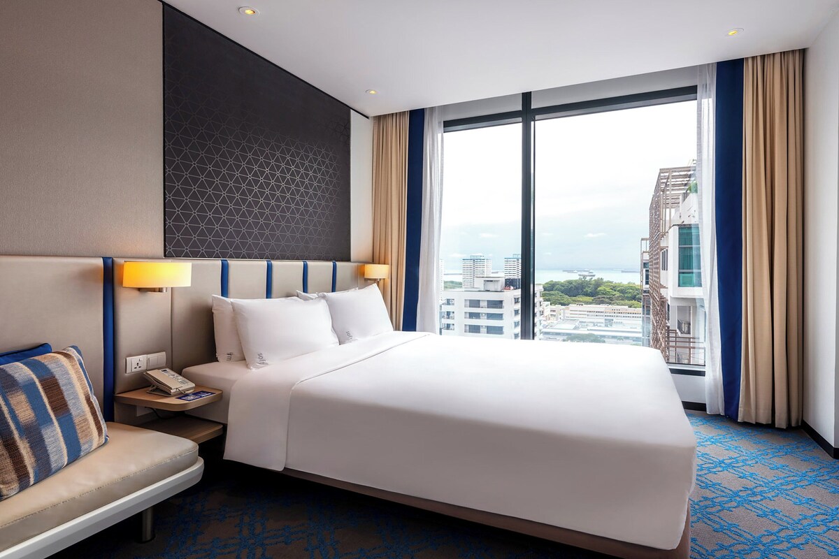 Ein Hotelzimmer mit einem großen Bett, einem Nachttisch mit Lampe, einem Sessel und Blick auf die Stadt durch große Fenster.