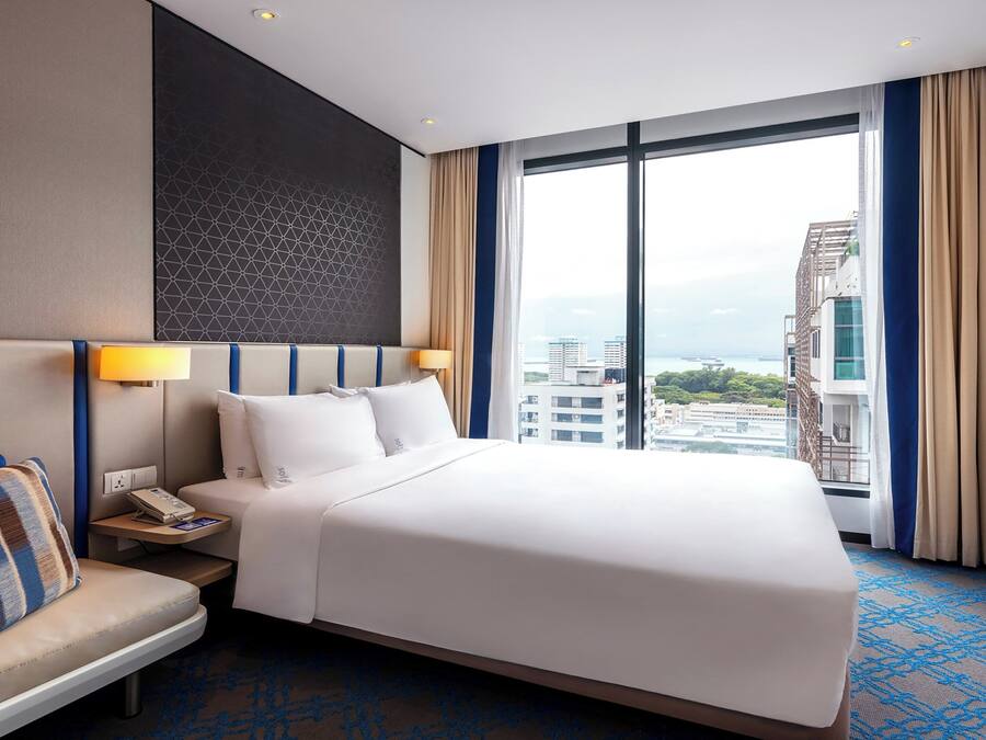 A hotel room with a large bed, a bedside table with a lamp, a seating area, and a view of the city through large windows.