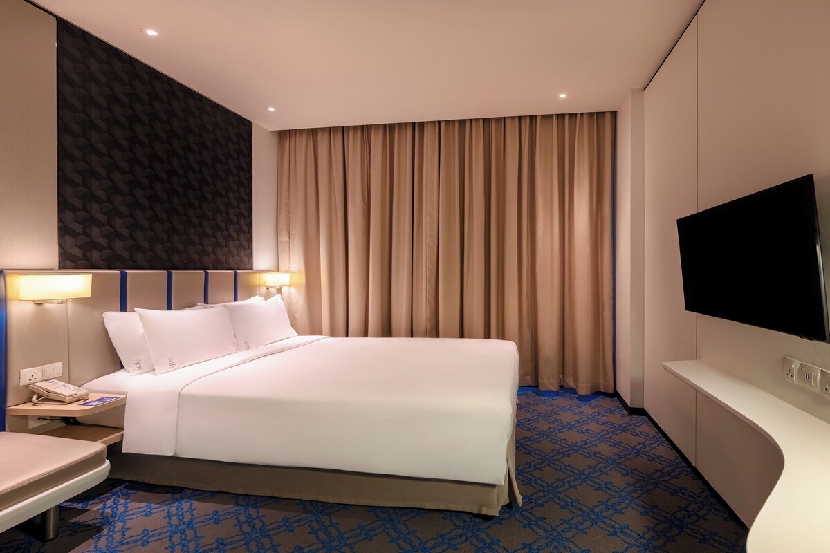 Ein Hotelzimmer mit einem großen Bett, einem Nachttisch mit Telefon, einem an der Wand montierten Flachbildfernseher und einem gemusterten Teppich.