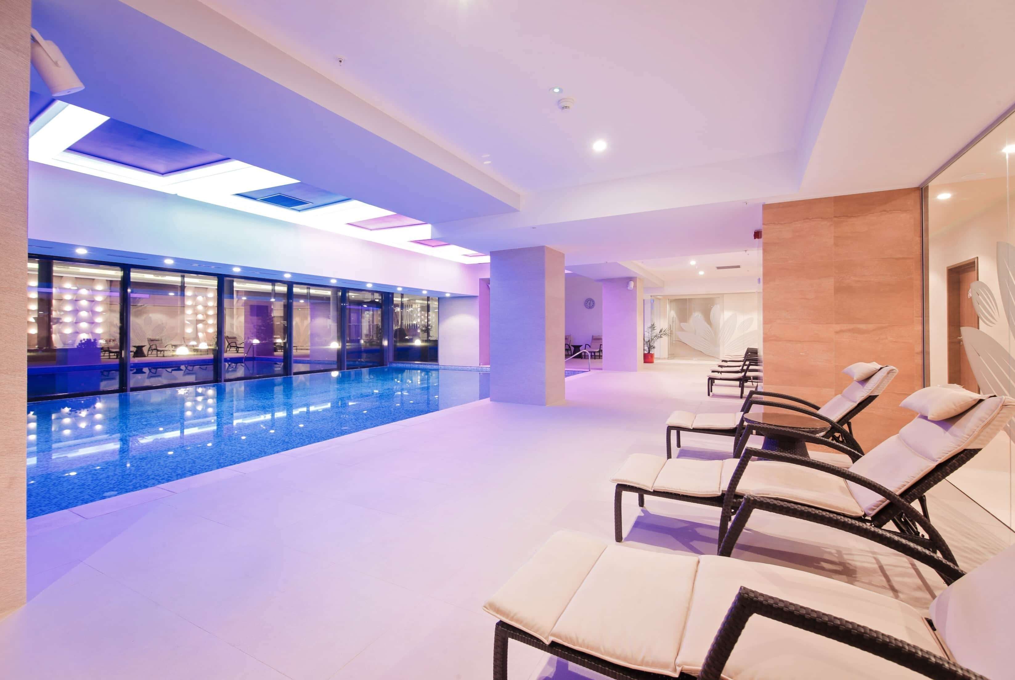 A modern indoor swimming pool area with lounge chairs and a glass wall.