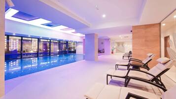 A modern indoor swimming pool area with lounge chairs and a glass wall.