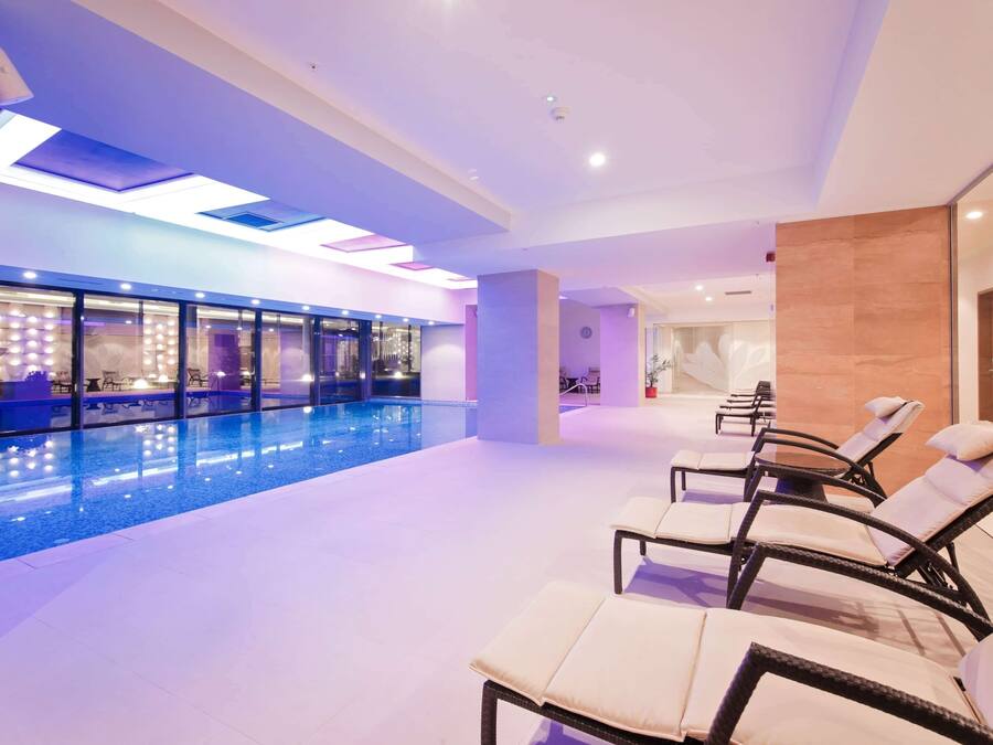 A modern indoor swimming pool area with lounge chairs and a glass wall.