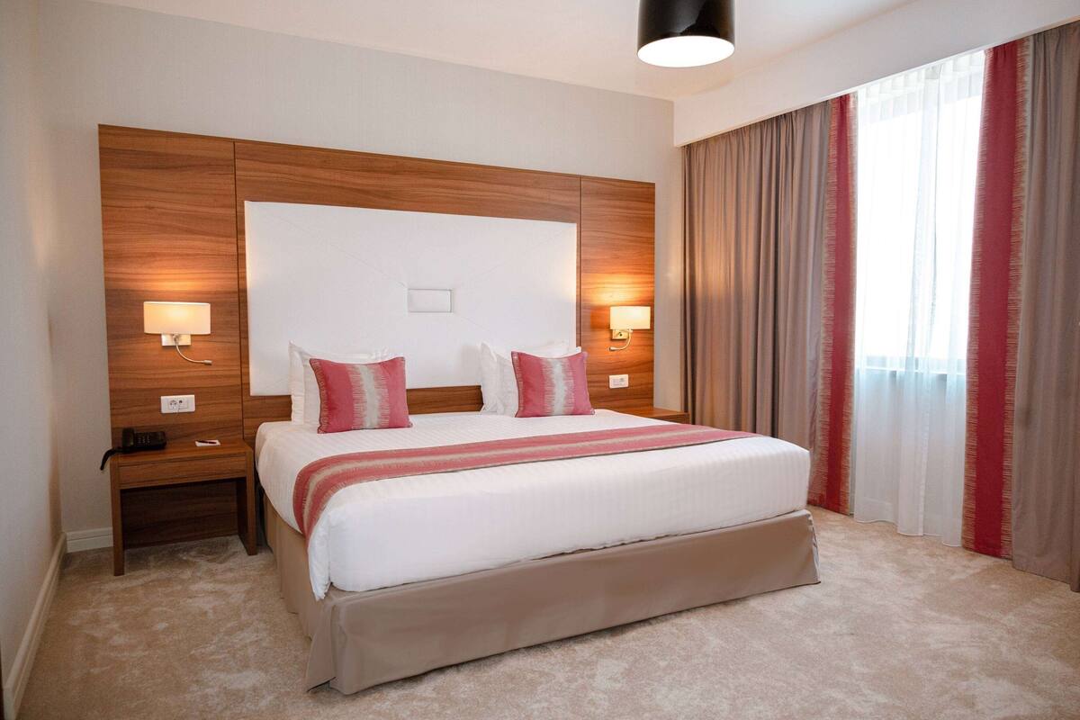 Una habitación de hotel con una cama grande, cabecera de madera, dos mesitas de noche y un pequeño mueble auxiliar.
