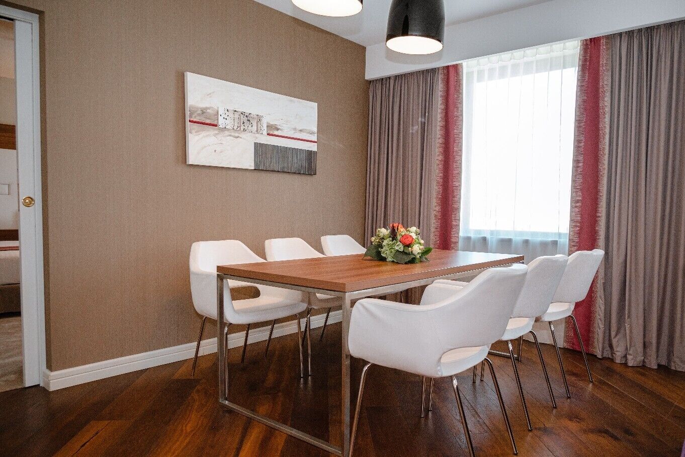 Een moderne hotelkamer met een houten eettafel, witte stoelen, een bloemdecoratie en een groot raam met gordijnen.