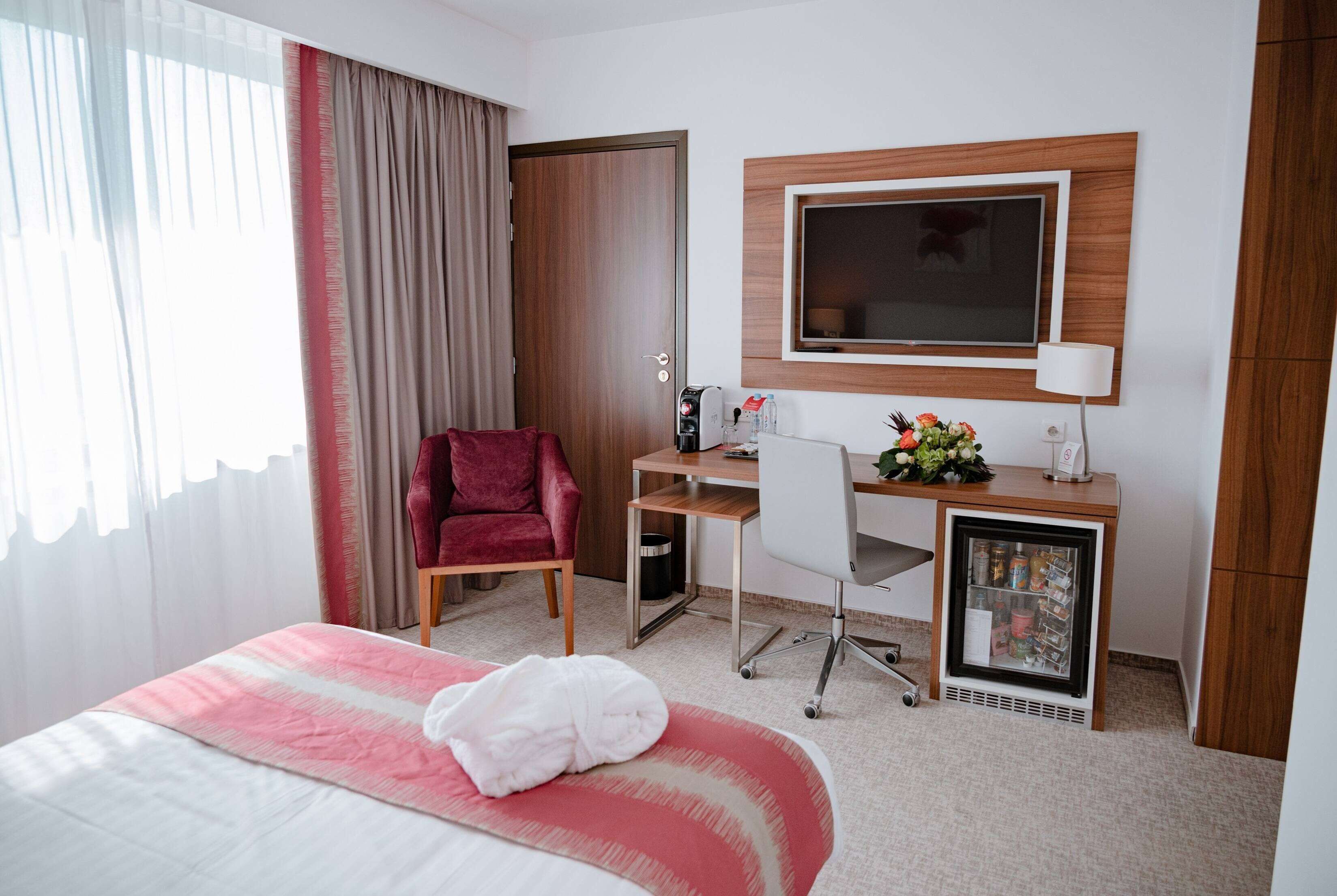 A hotel room with a bed, a desk with a computer, a TV, a mini-fridge, and a red chair.