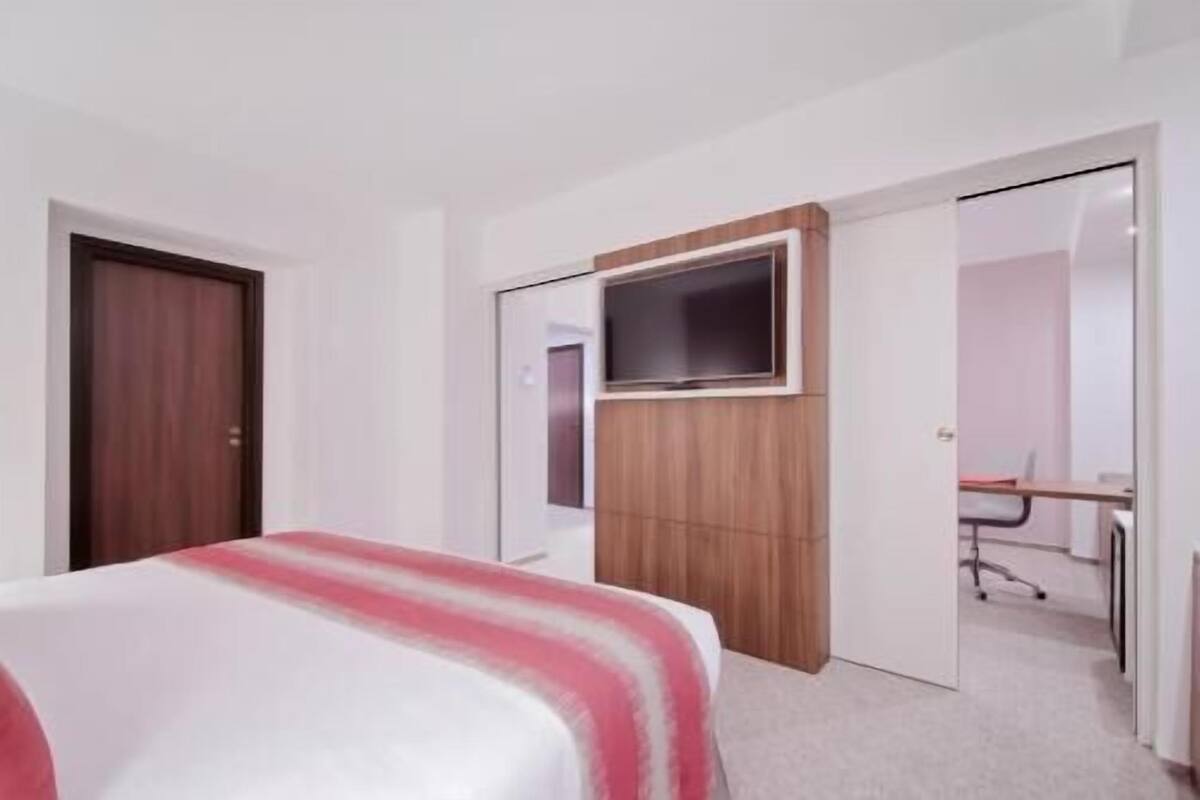 Una habitación de hotel con una cama, un televisor montado en un panel de madera, un escritorio con una silla y una puerta que conduce a otra habitación.