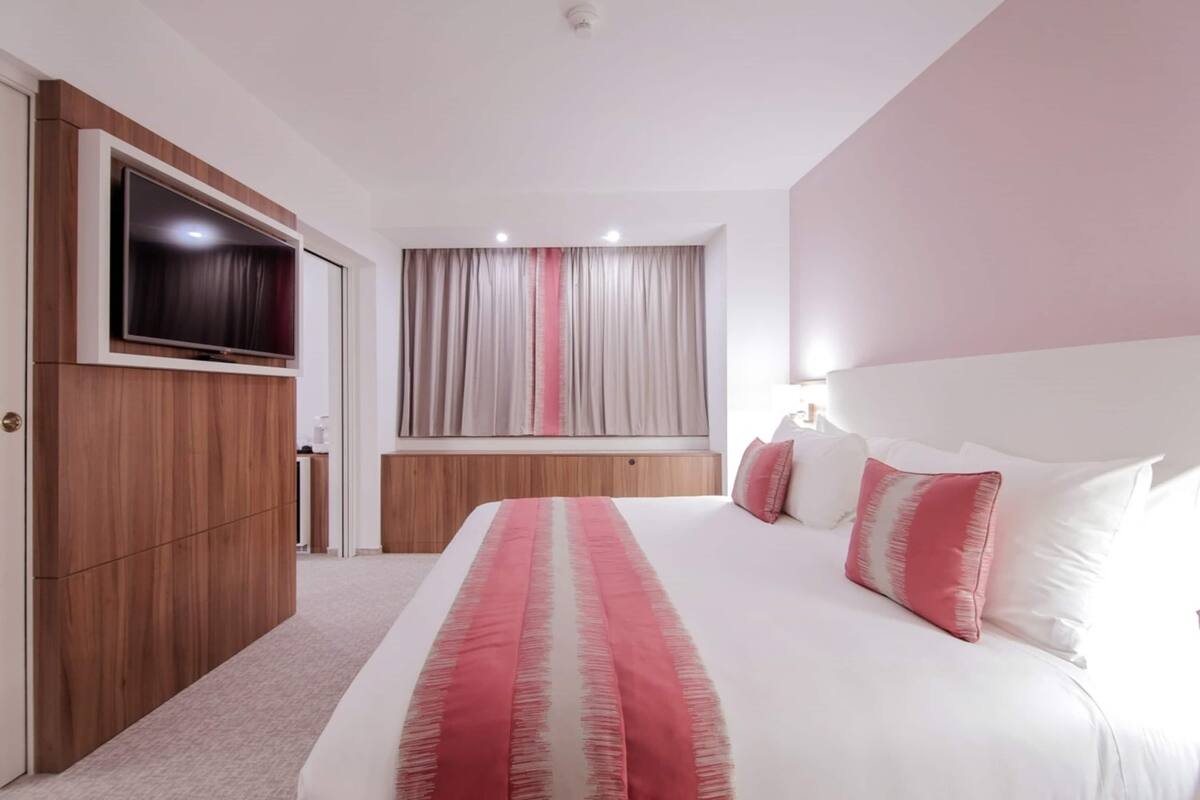Una habitación de hotel moderna con una cama grande, un televisor de pantalla plana montado en un panel de madera, un ventanal con cortinas y un cabecero con almohadas.