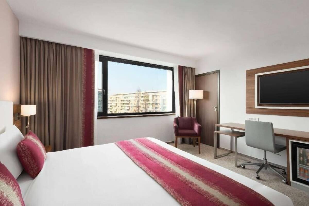 Habitación de hotel con cama, escritorio, silla y una ventana grande que ofrece vistas a la ciudad.