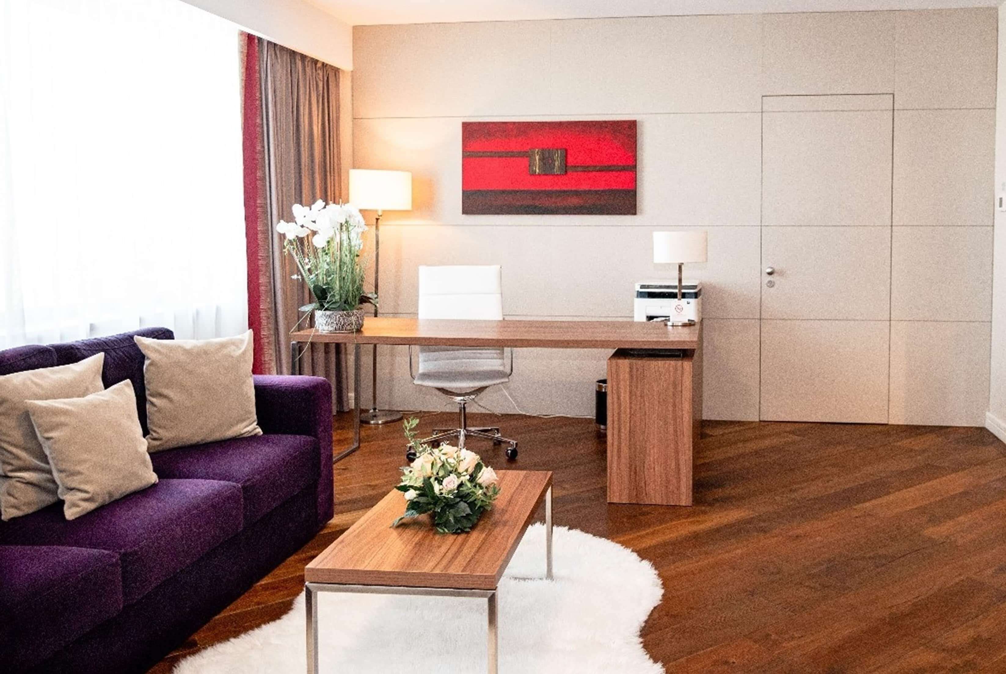 Une chambre d’hôtel moderne avec un canapé violet, une table basse en bois, un bureau avec un ordinateur et un tableau abstrait rouge accroché au mur.