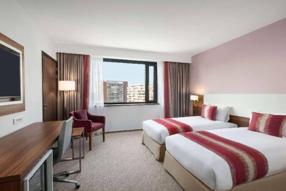 Habitación de hotel con dos camas, un escritorio, una silla, un televisor y una ventana con vista a la ciudad.