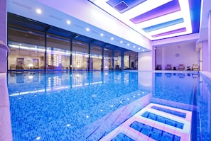 Una piscina interior moderna con paredes de vidrio transparente, suelo de baldosas e iluminación empotrada en el techo.