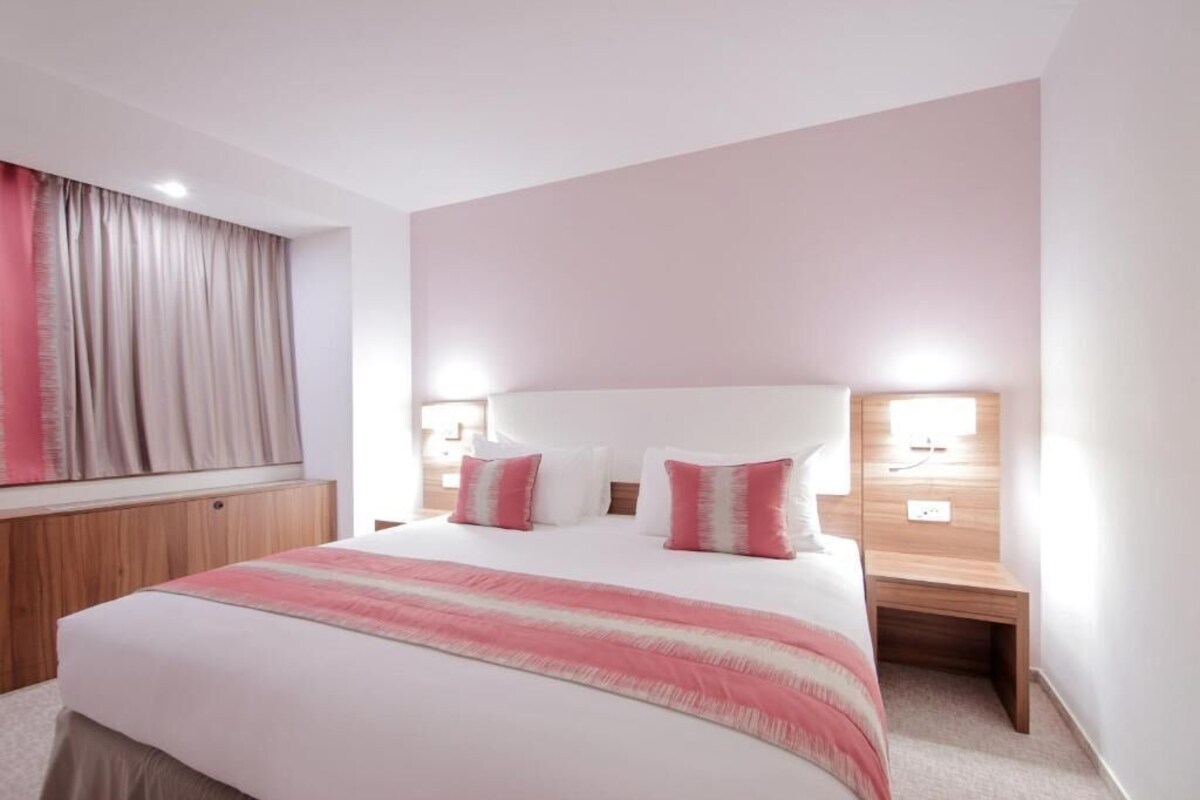 A hotel room with a large bed, two bedside tables, a headboard, and a window with curtains.