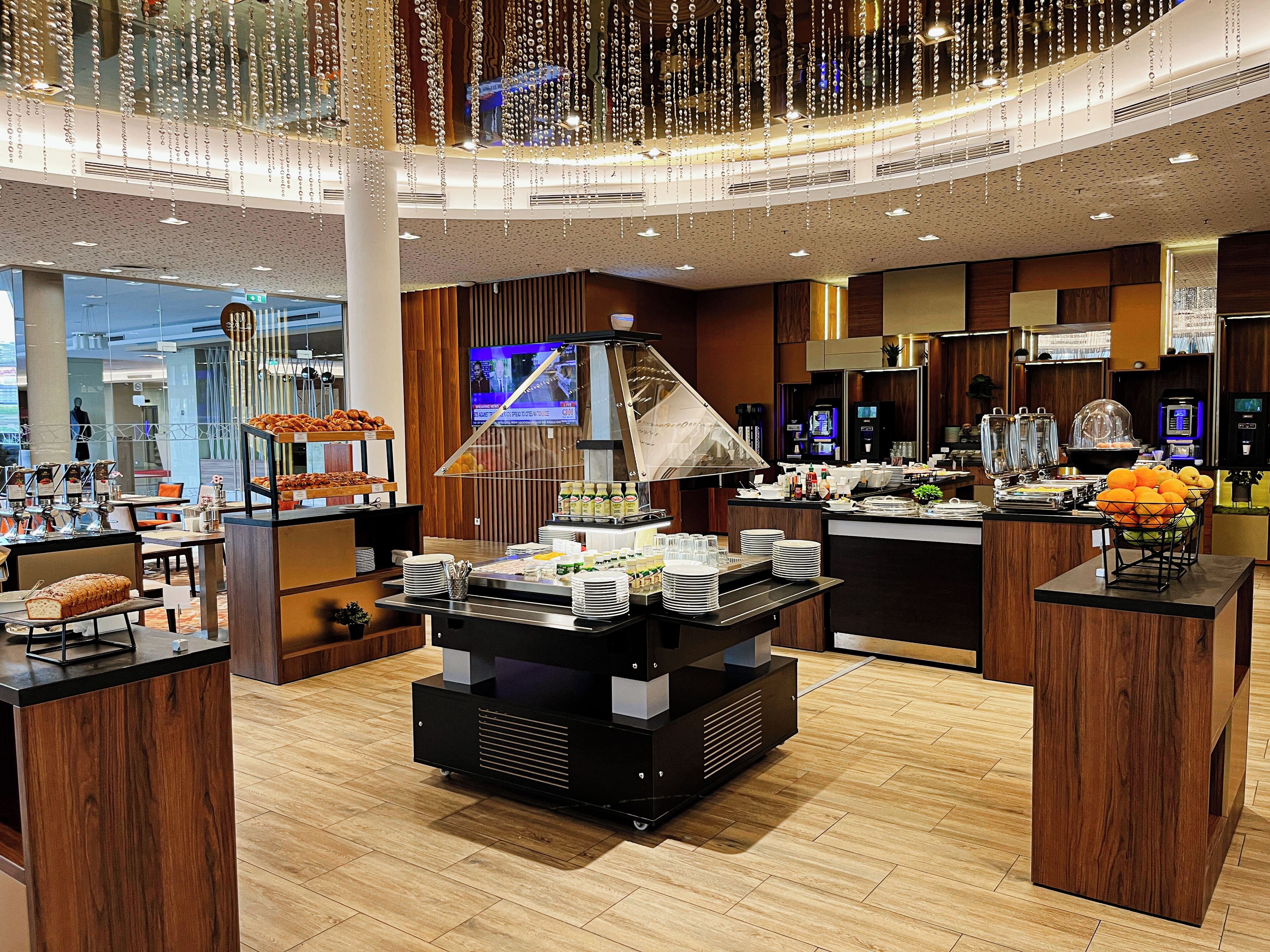 A modern hotel lobby with a buffet area, a television screen, and a coffee station.