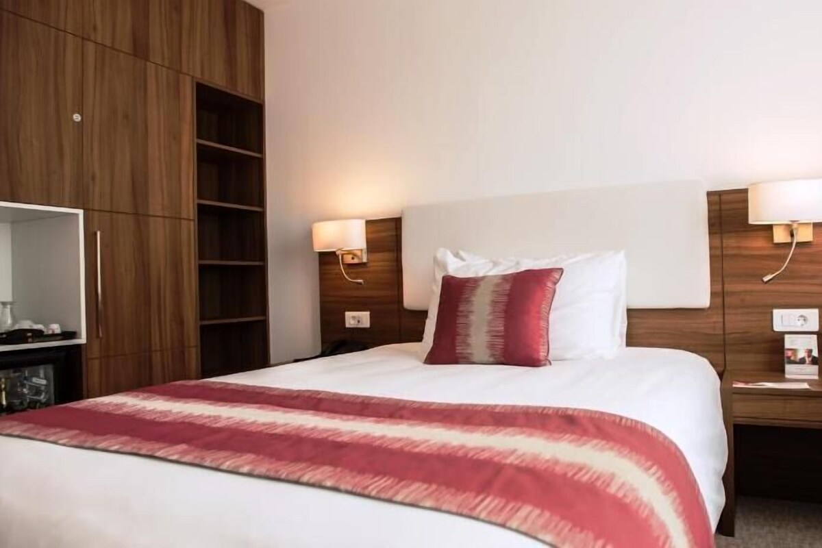 Una habitación de hotel con una cama grande, un armario, una mesita de noche con una lámpara y una pequeña mesa con cafetera.
