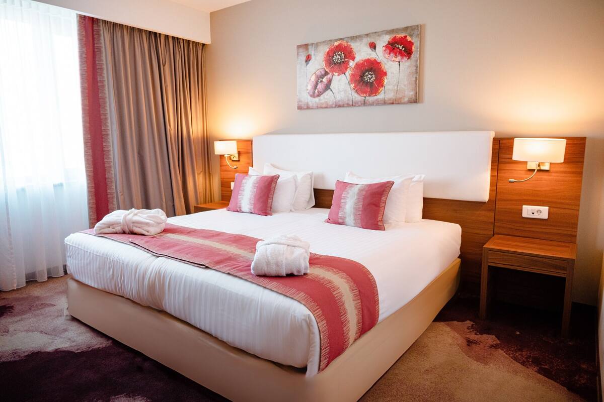 Habitación de hotel con una cama grande, dos mesitas de noche con lámparas, un cuadro en la pared y una ventana con cortinas.
