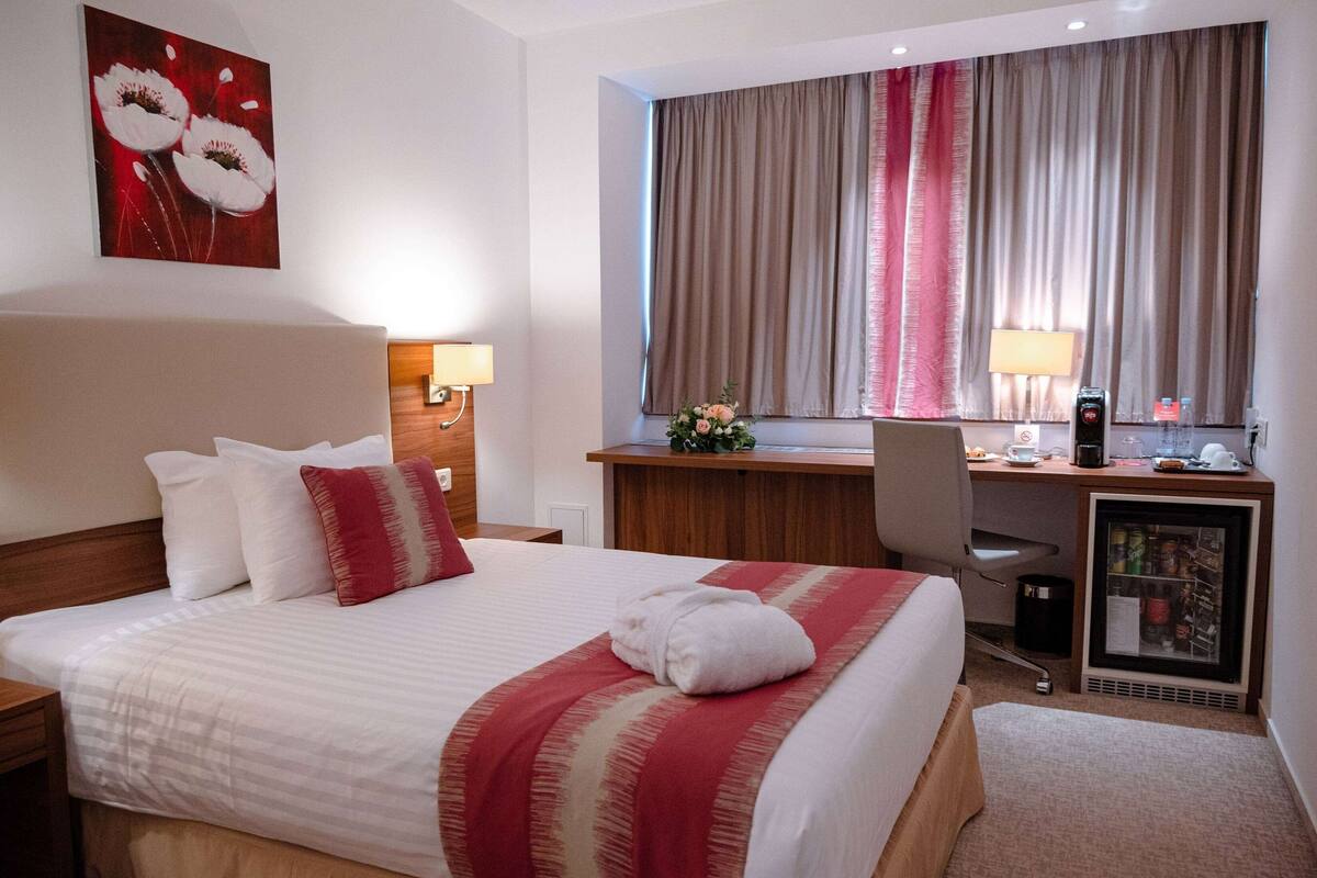 Una habitación de hotel con una cama grande, un escritorio, una silla, un refrigerador y una ventana con cortinas.