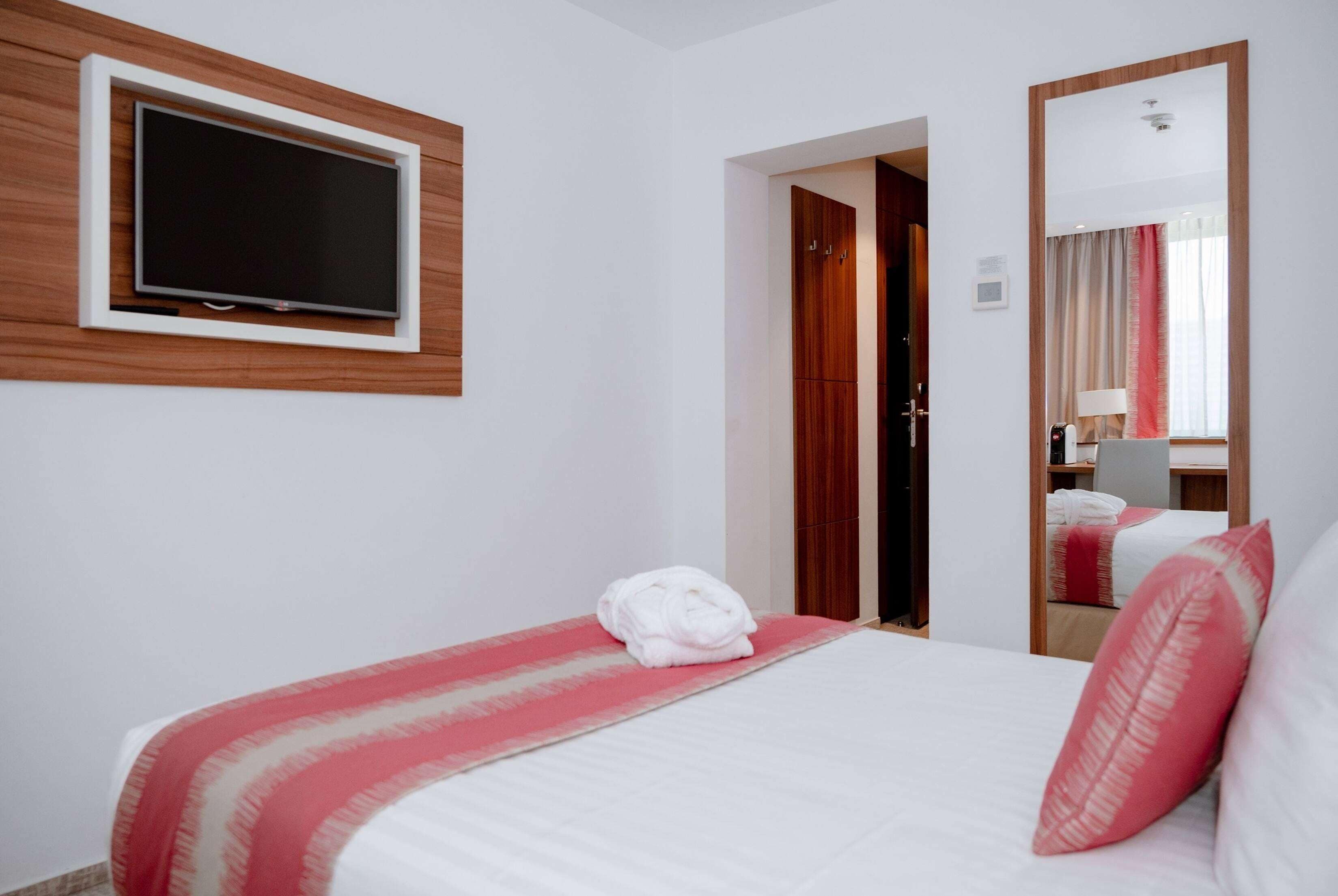 A hotel room with a bed, a TV mounted on the wall, a wardrobe, and a mirror reflecting part of the room.