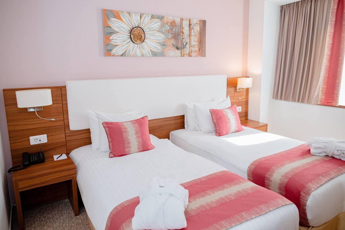 Habitación de hotel con dos camas, cada una con ropa blanca y detalles rosas, cabeceros de madera, lámparas de noche, un teléfono y un cuadro en la pared.
