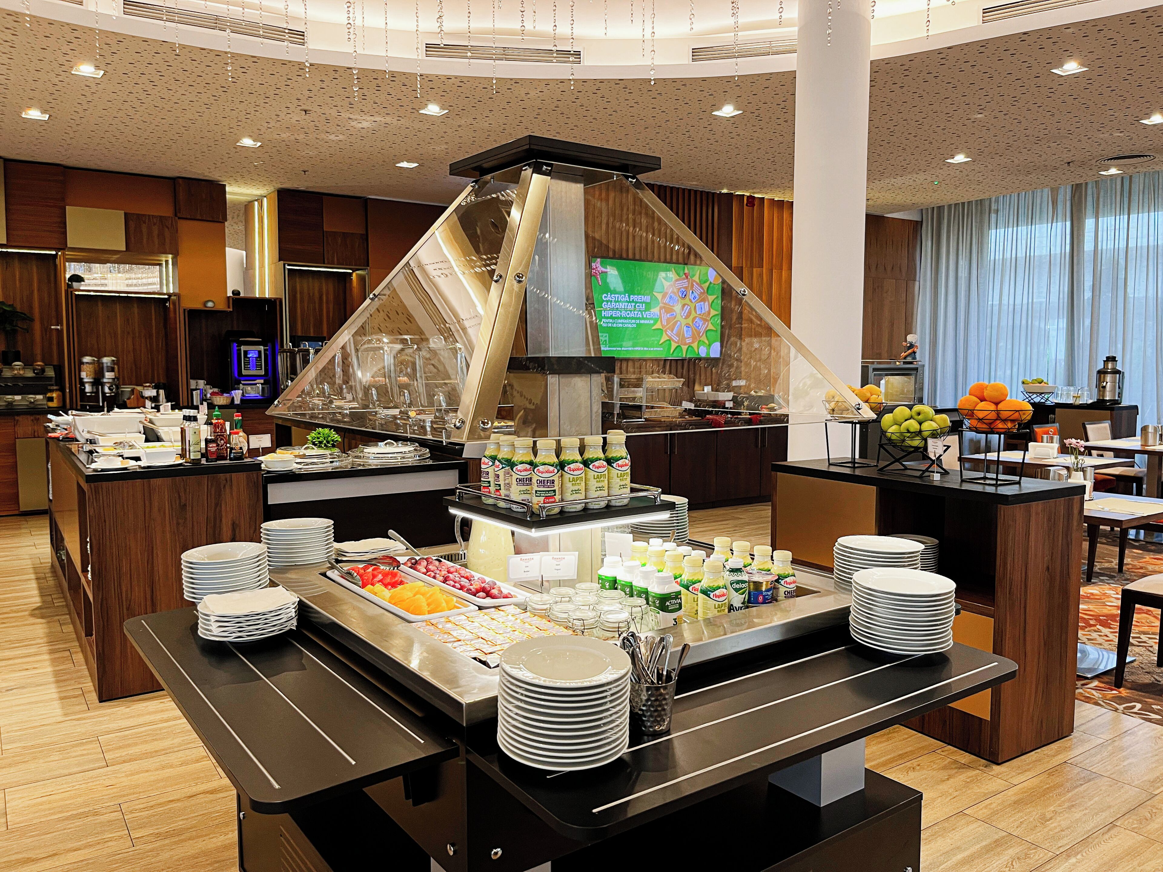 Buffet montado com diversos alimentos e bebidas, rodeado por mobiliário de madeira e um design moderno no teto.