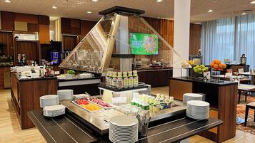 Buffet montado com diversos alimentos e bebidas, rodeado por mobiliário de madeira e um design moderno no teto.