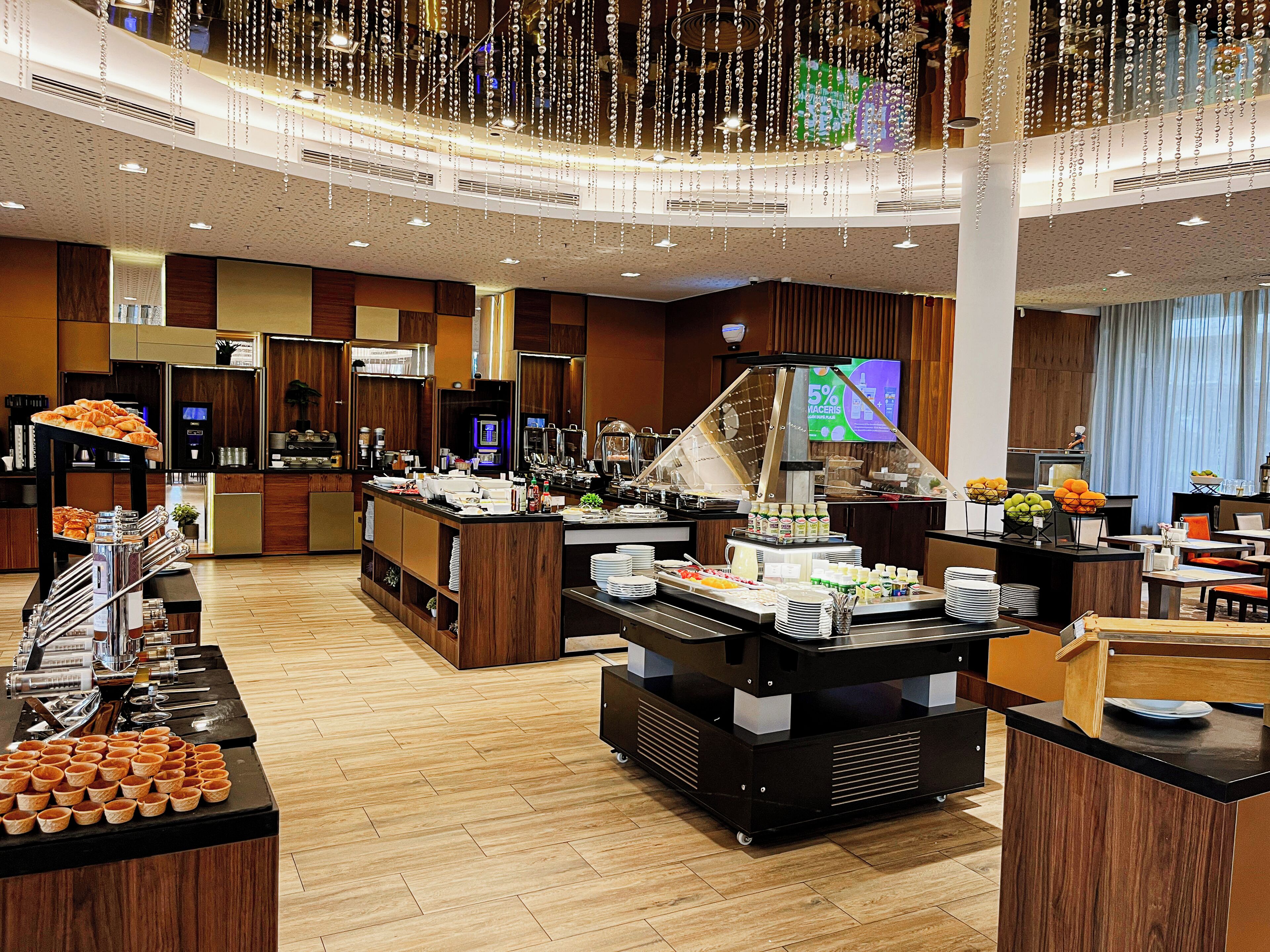 A hotel breakfast area with a buffet setup, a coffee station, and a television screen displaying a promotional offer.