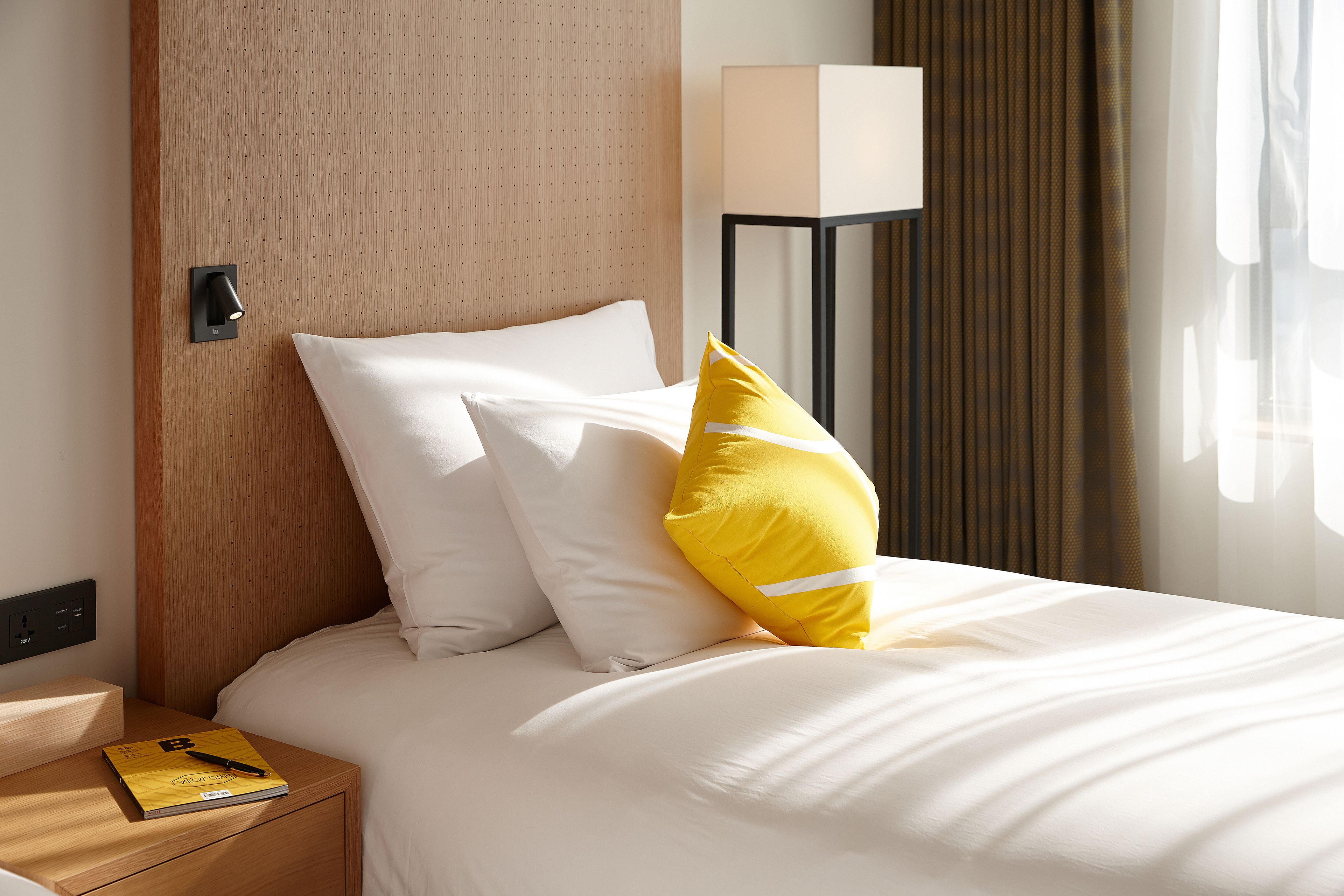 A neatly made bed with white and yellow pillows, a bedside table with a book, and a lamp.