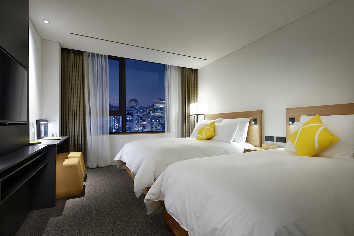 Een hotelkamer met twee bedden, een bureau en een groot raam met uitzicht op de stad.
