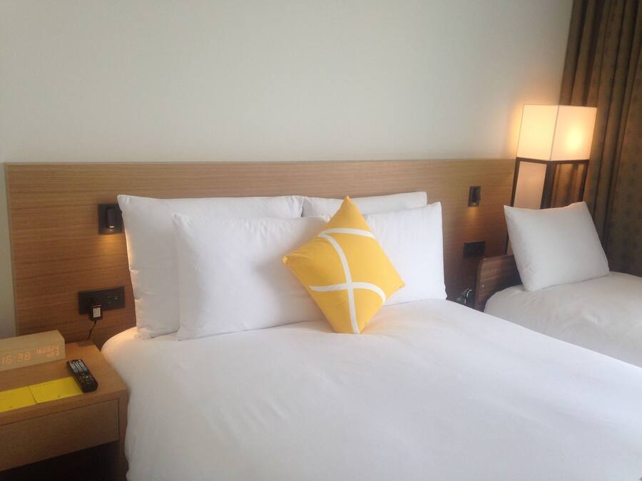 A hotel room with two beds, a nightstand with a clock, a remote control, and a lamp.
