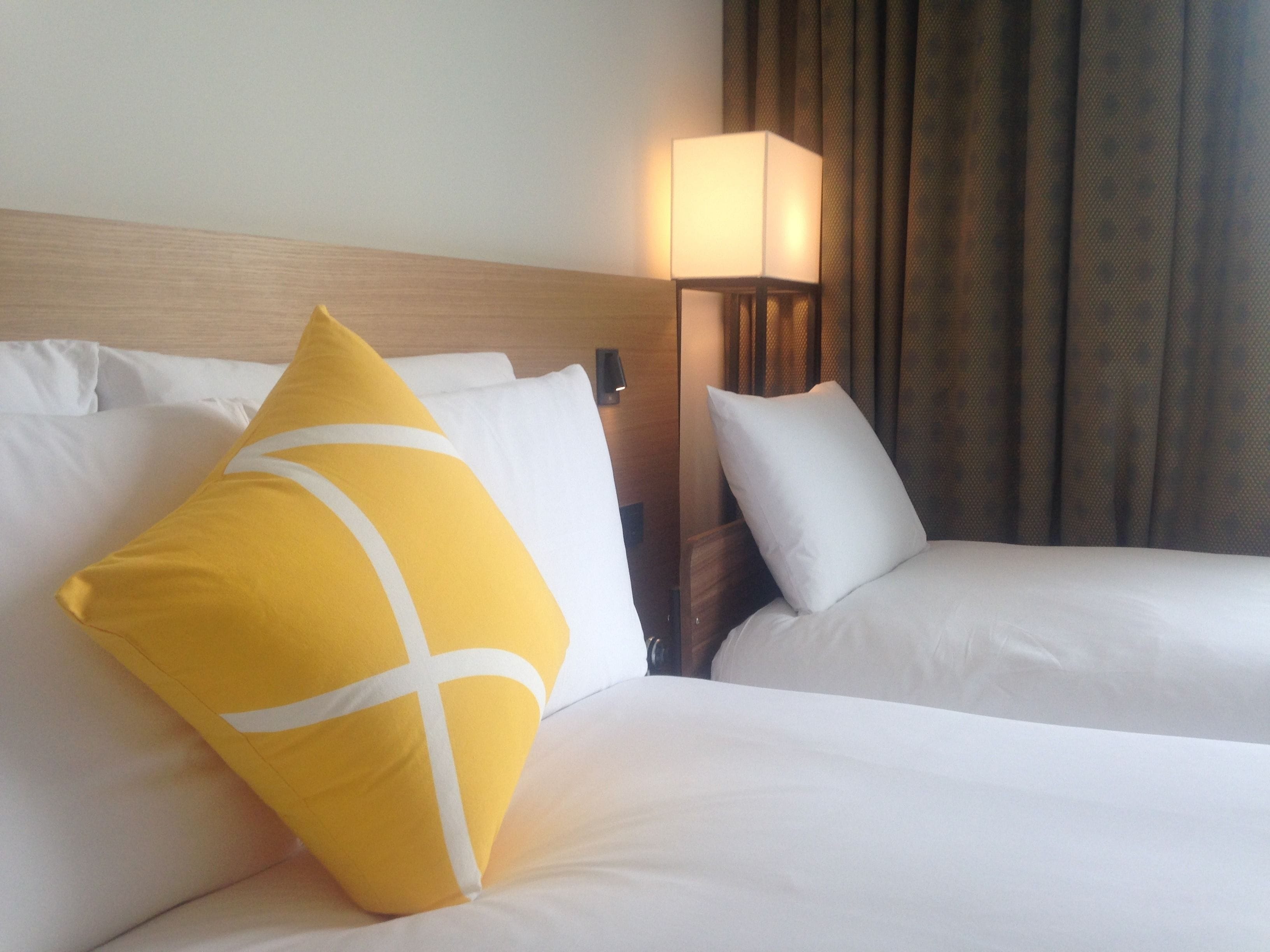 Une chambre d’hôtel avec deux lits, une tête de lit en bois, une table de chevet, une lampe et une fenêtre avec des rideaux.